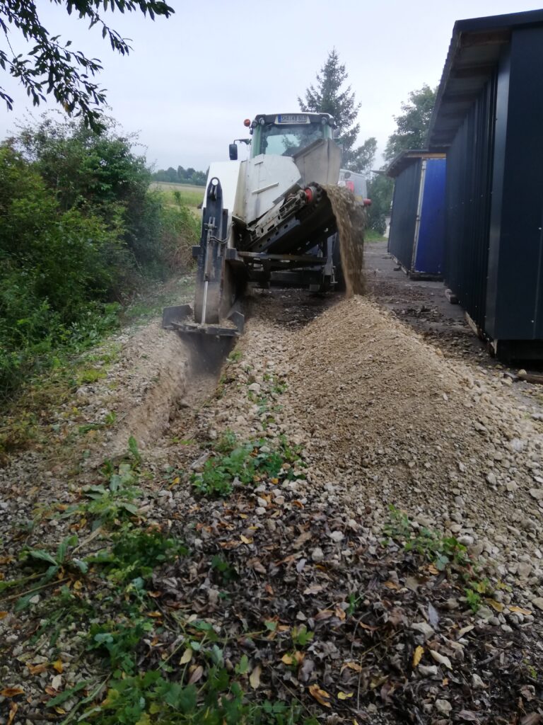 Ein Bagger bei Bauarbeiten auf einer Schotterstraße, umgeben von Baeumen und einem Gebaeude.