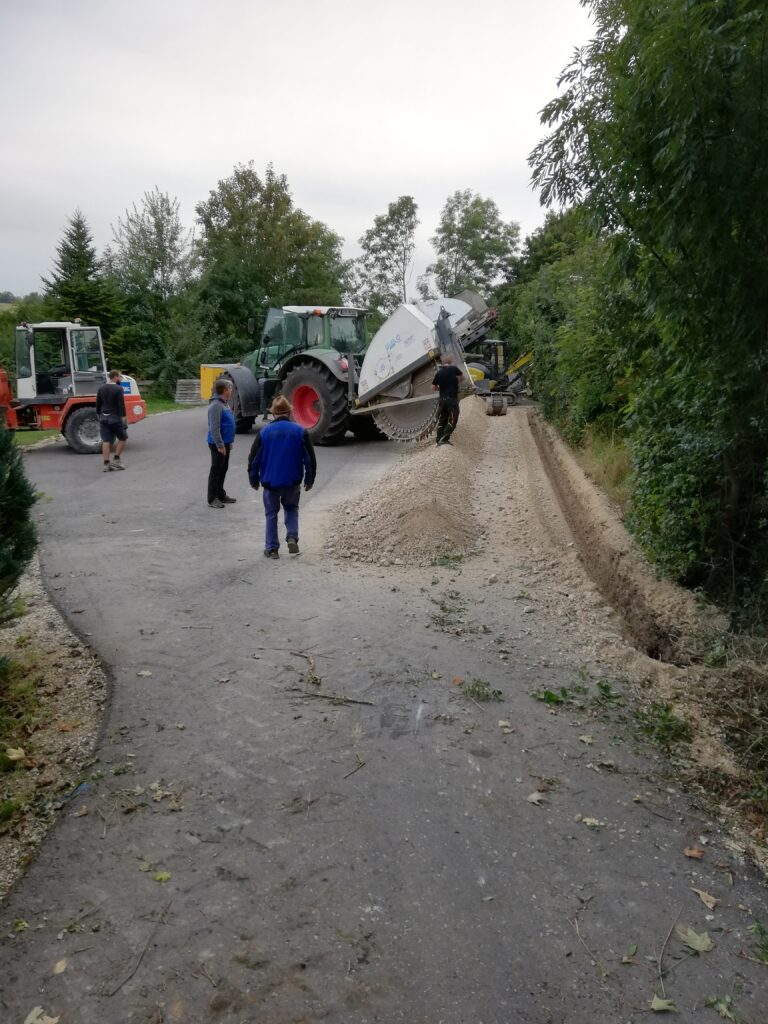 Menschen beobachten Bauarbeiten an einer Straße, umgeben von Baeumen.