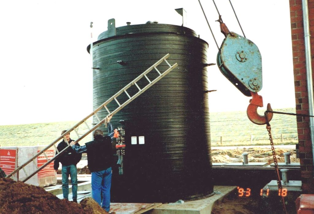 Großer zylindrischer Behaelter wird von einem Kran gehoben, waehrend Menschen daneben stehen, in den Anfangsjahren des Familienunternehmens Ebert Kunststoffbearbeitung.